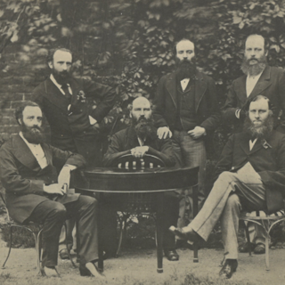 Black and white photo of six bearded men gathered around a table dressed in formal suits from the 1800s, threee seated and three standing