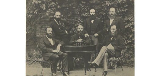 Black and white photo of six bearded men gathered around a table dressed in formal suits from the 1800s, threee seated and three standing