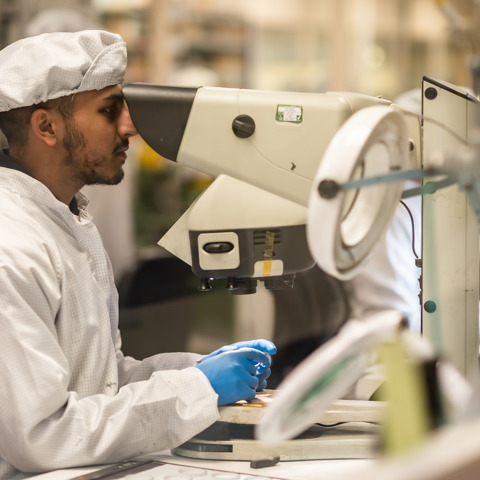 Male scientist looking through large microscope, wearing white overalls and white hat, in factory clean room